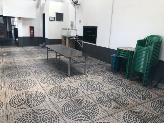 Salão de festas com mesas, cadeiras empilhadas e churrasqueira